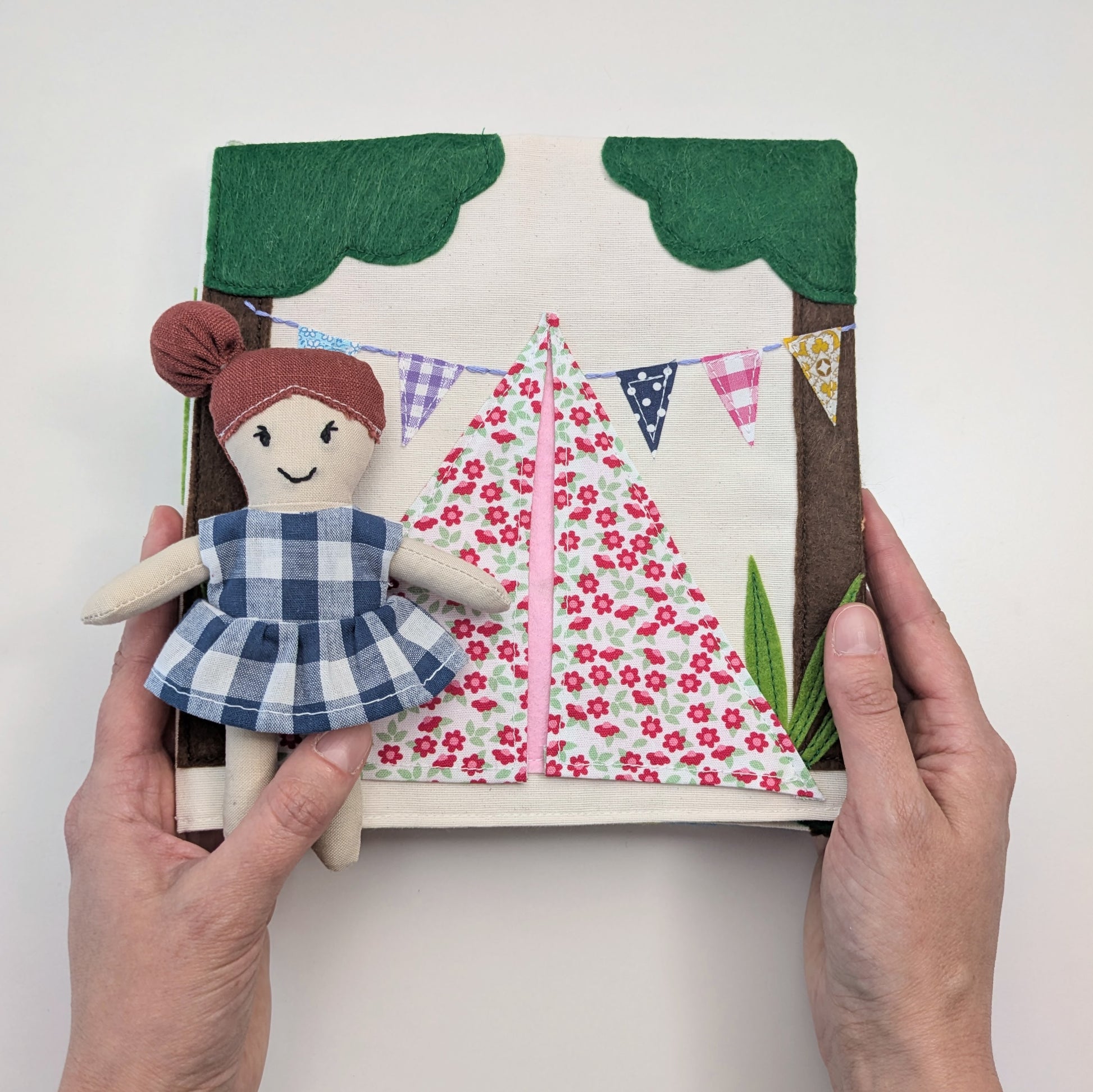 Handmade doll with a checkered dress held in front of a fabric scene with a sailboat and bunting.