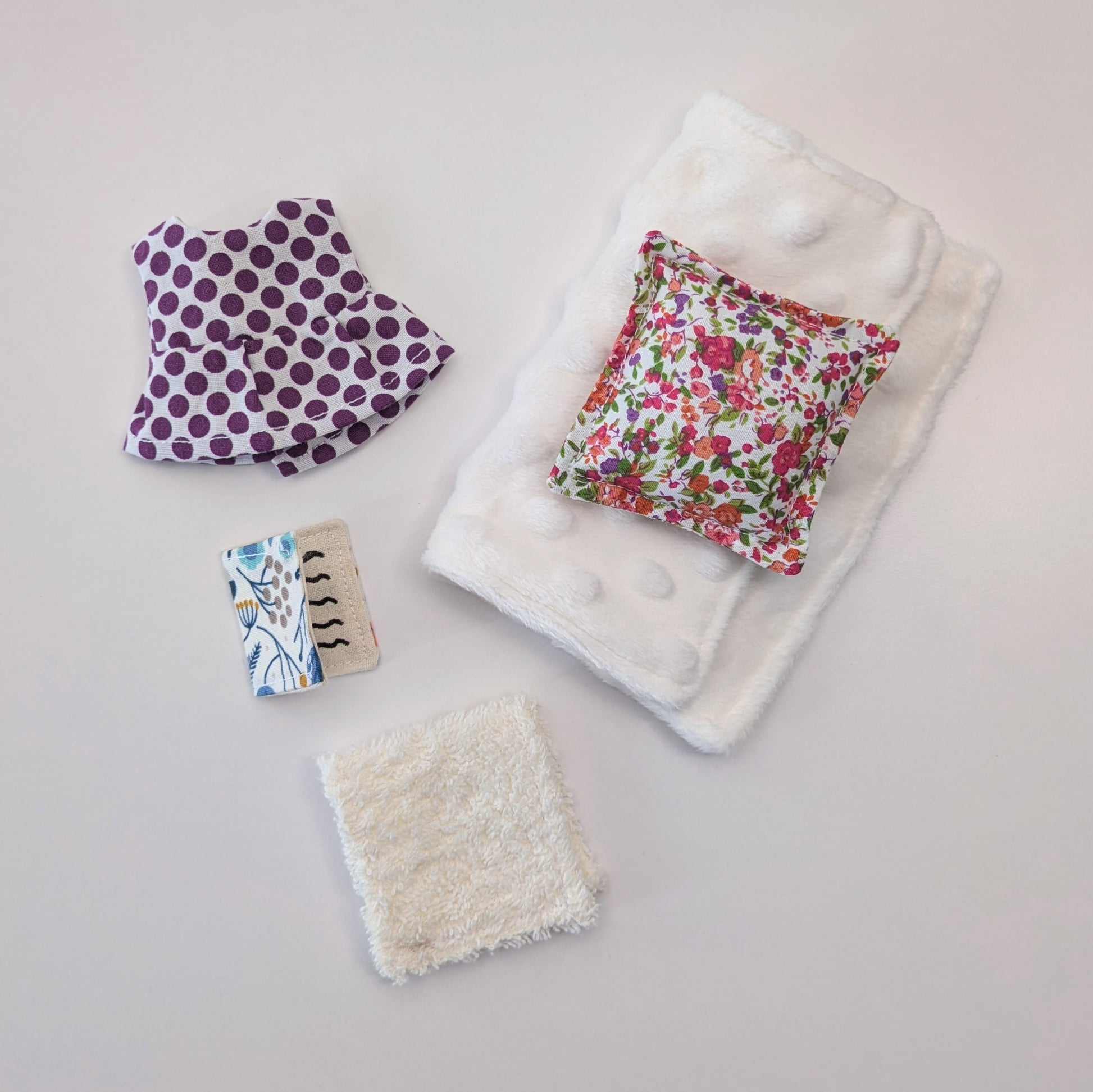 Collection of baby items including a floral-patterned blanket, a purple polka dot item, and a textured white square on a light gray background.